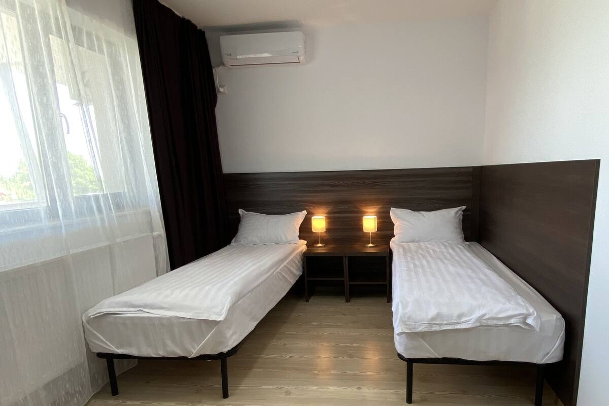Habitació amb dos llits individuals, aire condicionat fixat al sostre i una finestra amb cortines translúcides.