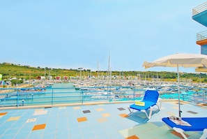 Un espace piscine avec des chaises longues bleues et des parasols, entouré d’une marina où sont amarrés de nombreux bateaux.