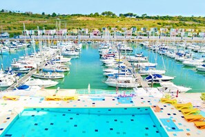 Un puerto deportivo con numerosas embarcaciones amarradas en aguas tranquilas, una piscina con tumbonas y una colina al fondo.