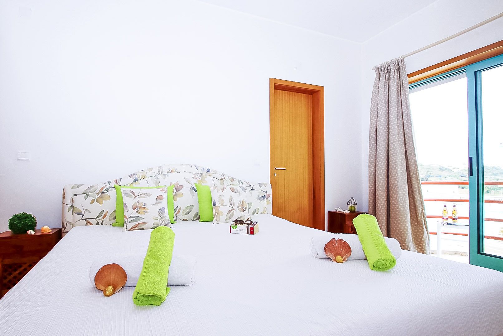 A hotel room with a bed, a wooden door, a window with curtains, and a small table with a plant.