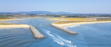 Une vue aérienne d’une zone côtière, avec un ciel dégagé, une plage de sable et une voie navigable où l’on peut apercevoir des bateaux.