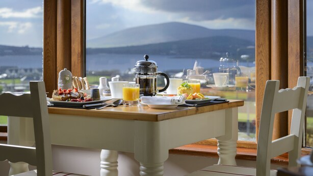 A breakfast table with a coffee pot, plates, glasses, and a view of a lake and mountains. - Greenmount House (Dingle)