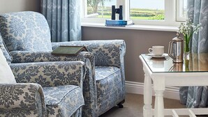 A room with a large window, a blue patterned sofa, a small table with a cup and a book, and a view of a lake and greenery. - Greenmount House (Dingle)