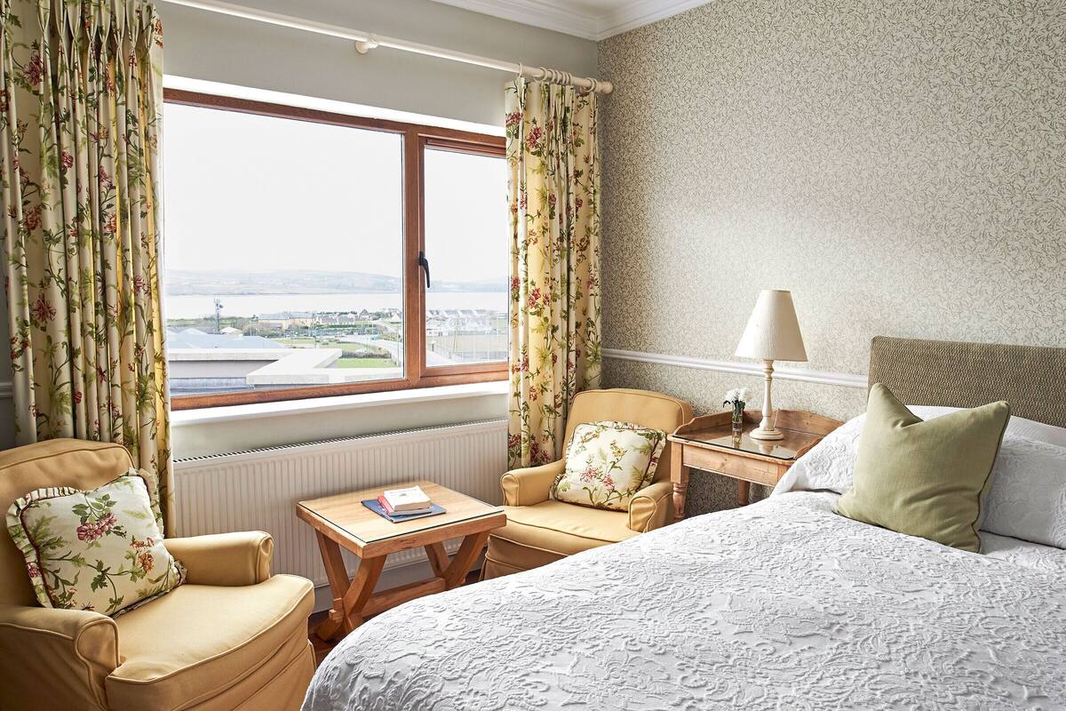 Habitación de hotel con cama, dos sillones, una mesita, una lámpara y vistas a un paisaje urbano a través de un gran ventanal.