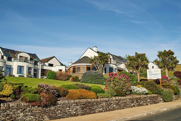 Une rangée de maisons modernes à deux étages, avec un jardin aménagé à l’avant, un mur en pierre et une enseigne indiquant « Greenmount Houses ».