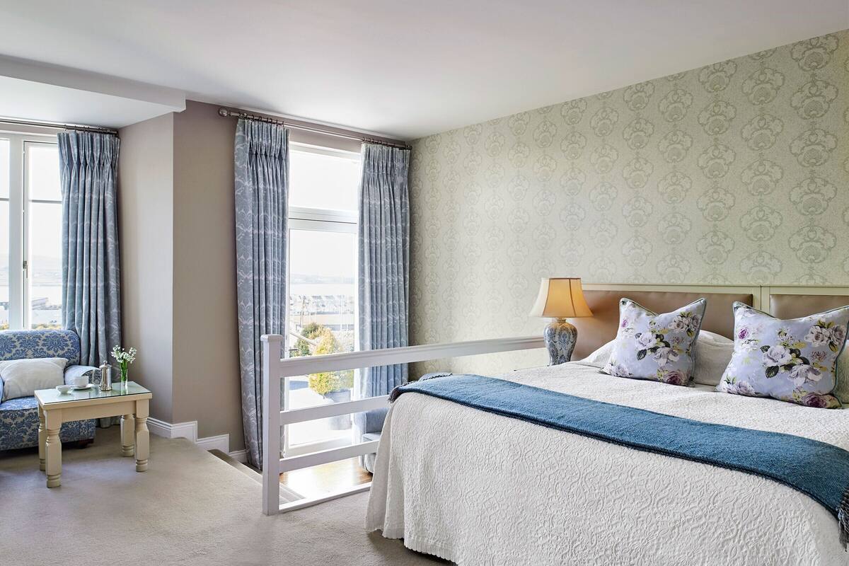 Un dormitorio con una cama, una mesita de noche, una ventana con cortinas y un papel tapiz estampado.