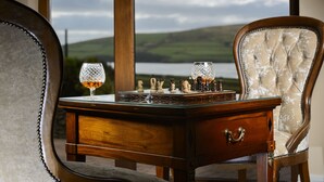 A wooden desk with a chess set and two crystal glasses, positioned in front of a window with a scenic view of hills and water. - Greenmount House (Dingle)