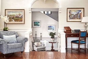 A room with a wooden floor, a grey patterned armchair, a small white chair, a wooden desk with a chair, and a staircase leading to another room.