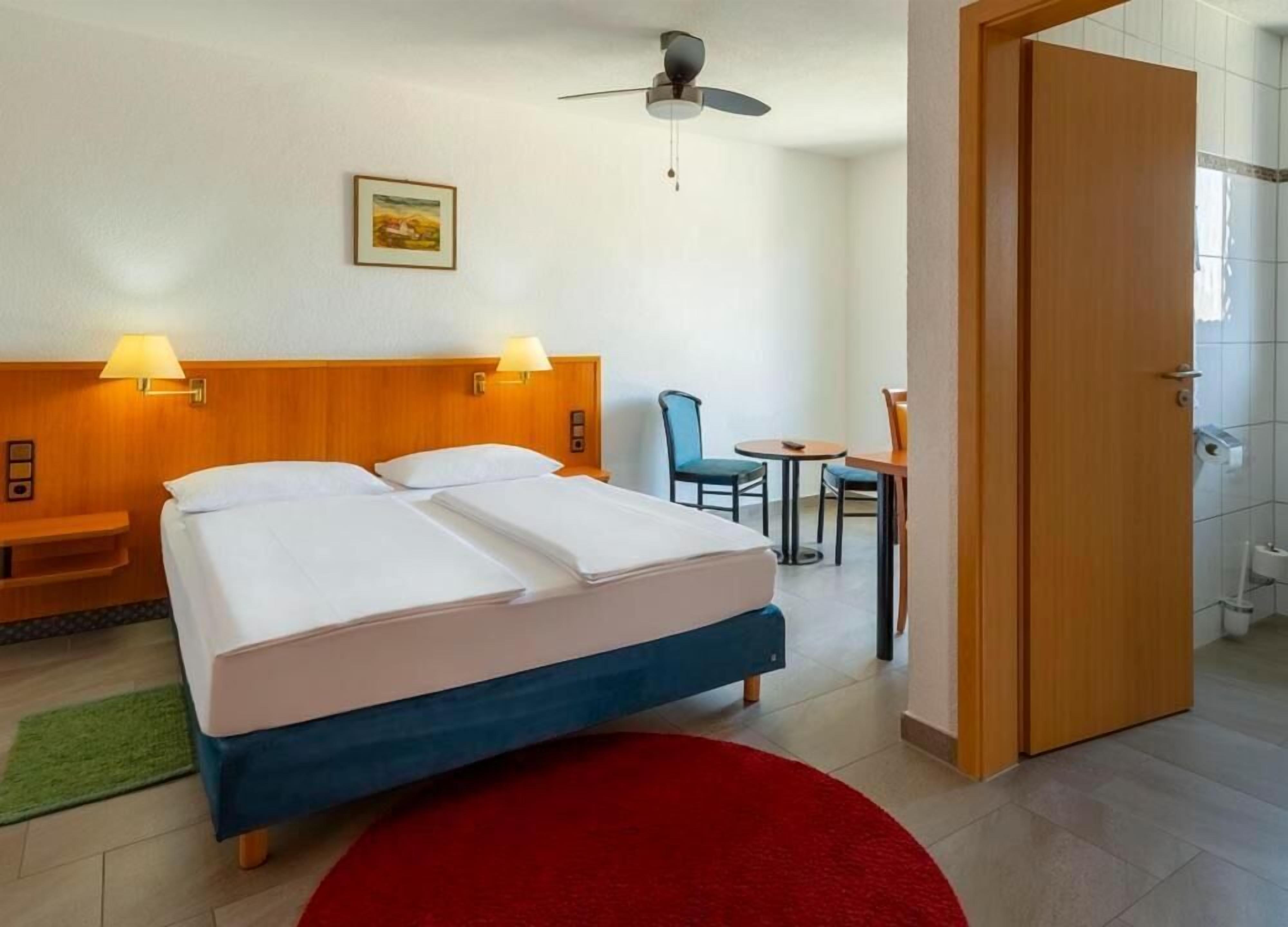 Habitación de hotel con cama doble, dos lámparas de noche, ventilador de techo, una pequeña mesa con sillas y una alfombra roja.