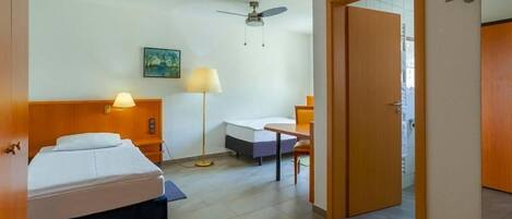 A hotel room with two beds, a desk, a chair, a ceiling fan, and a door leading to another room.