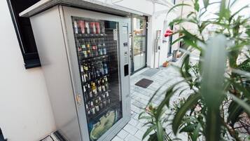 Distributore automatico con diverse bevande e snack, situato all'esterno di un edificio con pavimento in piastrelle e una pianta in vaso nelle vicinanze.