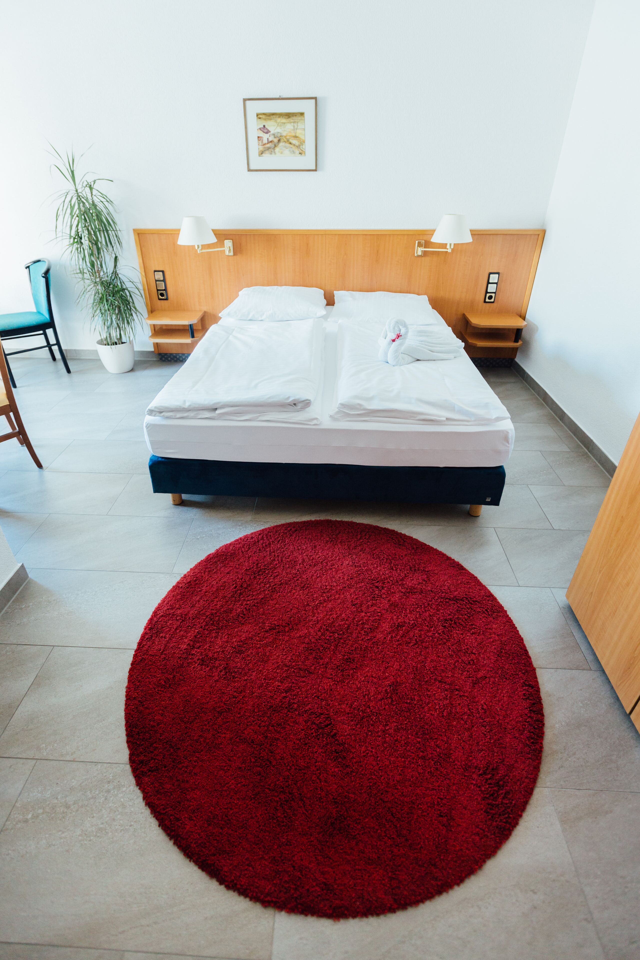 A neatly made bed with white linens, a red circular rug in the foreground, and a wooden headboard with wall-mounted lamps.