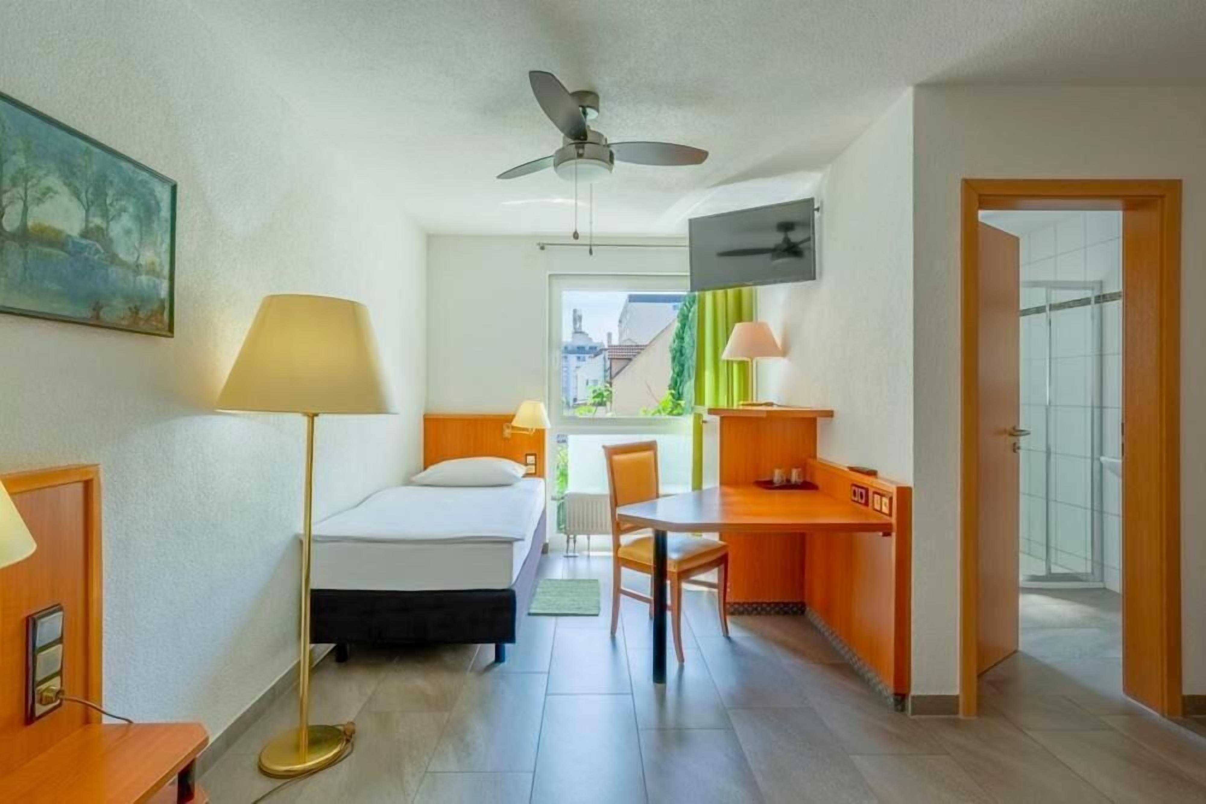 Una camera d'albergo con un letto, una scrivania, una sedia, una lampada, una ventola a soffitto e un quadro appeso al muro.