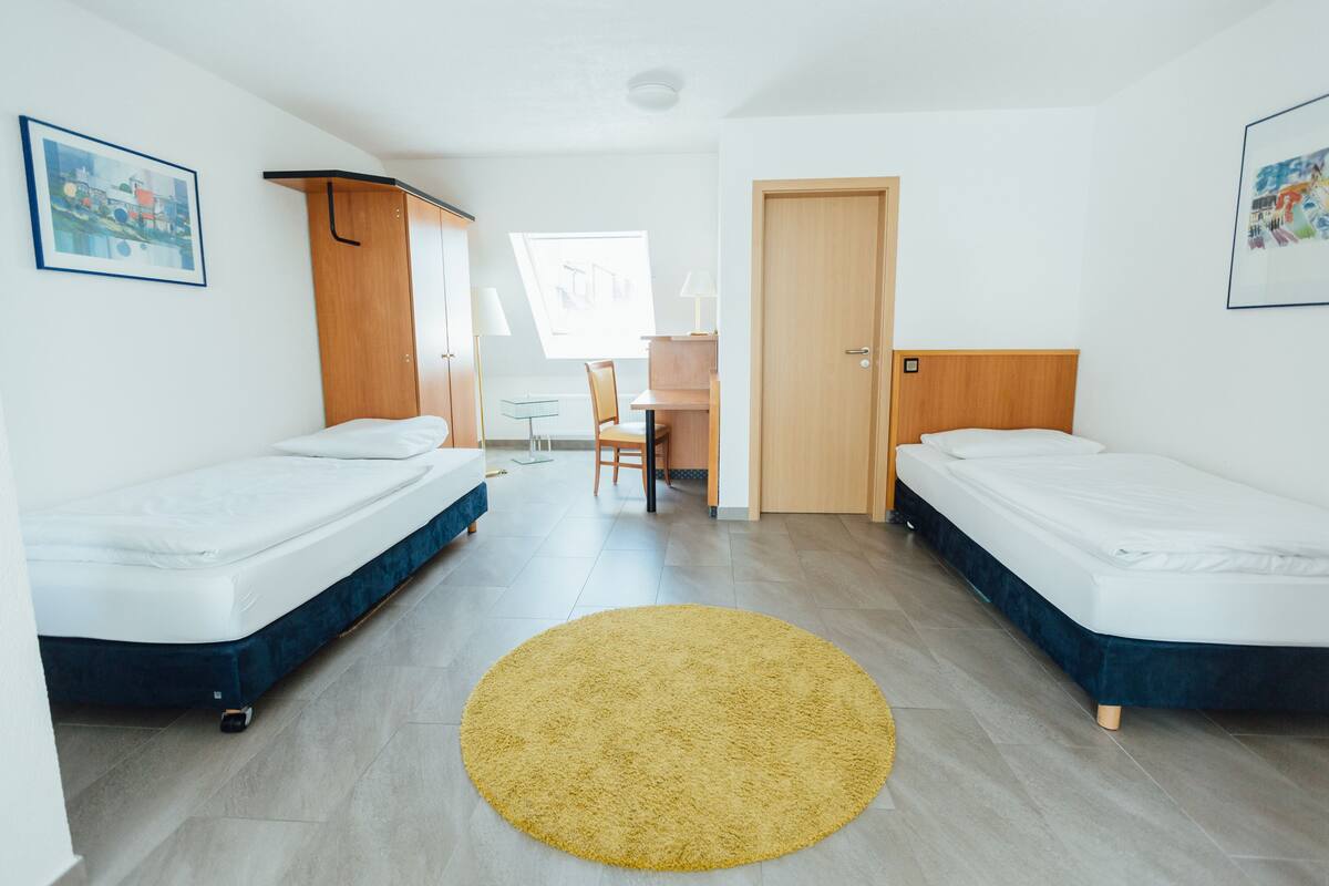 Une chambre moderne et bien éclairée, équipée de deux lits simples, d’un petit bureau et d’une chaise.