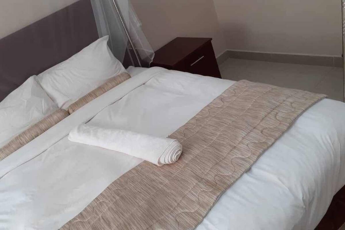 A neatly made bed with white linens and a beige quilt, a wooden nightstand beside it, and a plain wall in the background.
