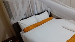 A neatly made bed with white and orange bedding, a dark wooden nightstand, and a white curtain. - Delight Lodge (Blantyre)