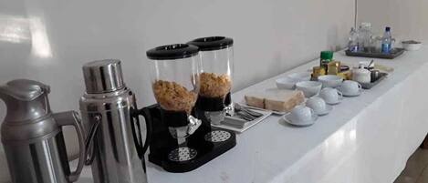 Mesa blanca con dispensadores de café, tazones y cubiertos en una barra tipo buffet.
