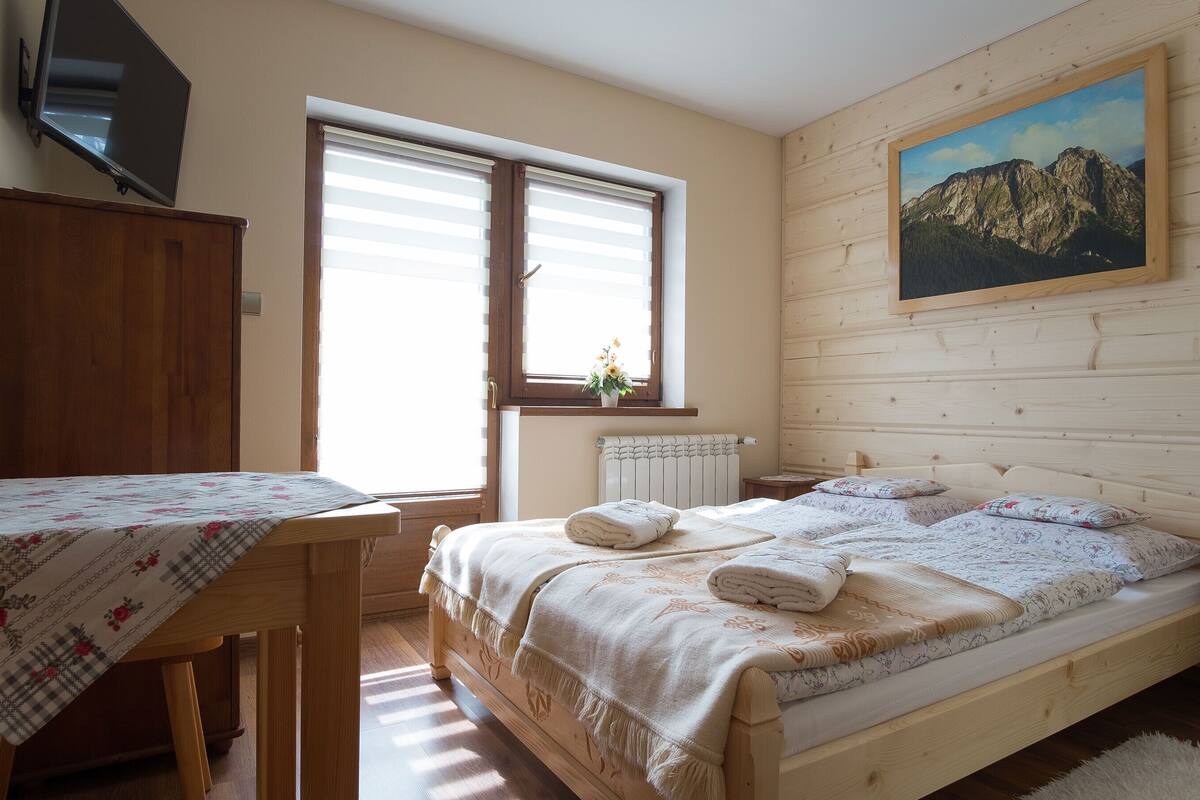 A bedroom with a wooden bed, a desk, a radiator, and a window with blinds.
