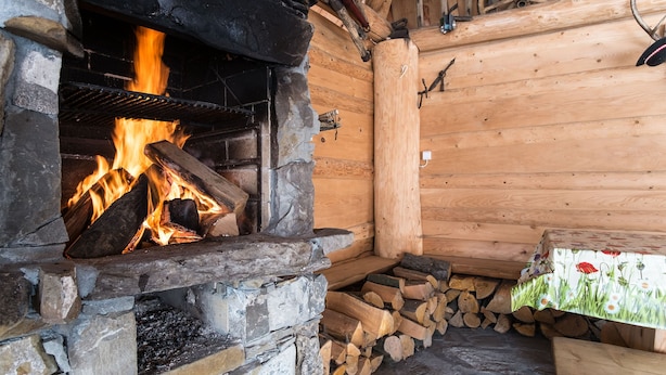 Una chimenea de piedra con fuego encendido en su interior, leña apilada a un lado y un interior de madera rústica.