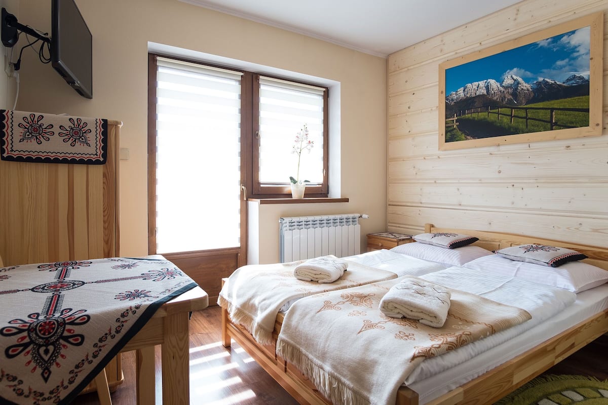 A bedroom with a wooden bed, a window with blinds, a radiator, a table with a patterned tablecloth, and a painting on the wall.