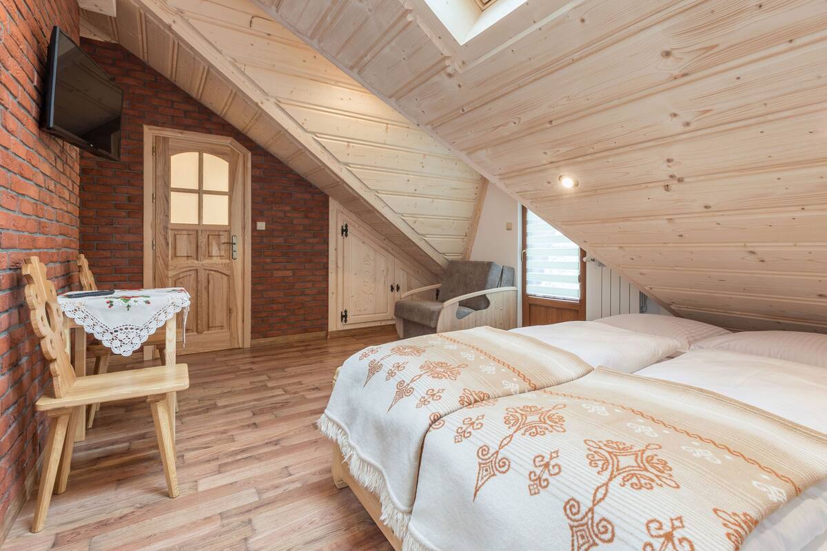 A cozy attic bedroom with a wooden ceiling, a bed with patterned bedding, a wooden chair, and a table with a lace tablecloth.
