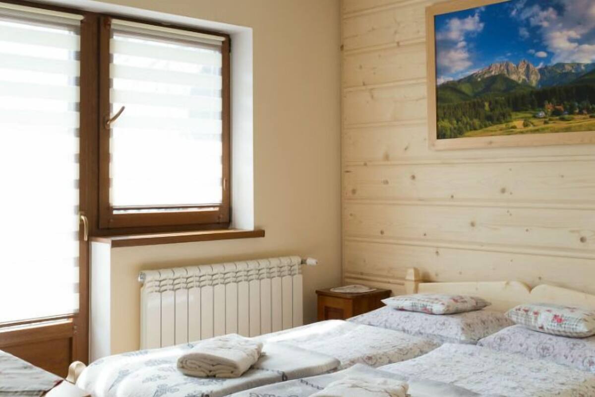 A bedroom with a bed, a window, a radiator, a nightstand, and a painting of mountains.