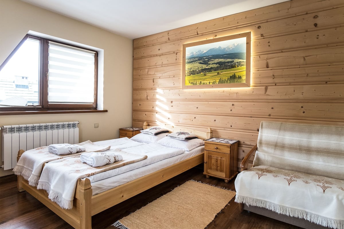 A bedroom with a wooden bed, a nightstand, a chair, a window with blinds, and a picture on the wall.