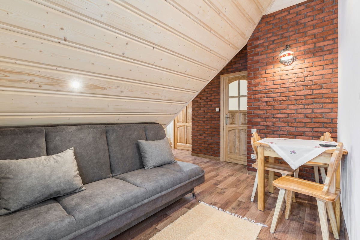 A cozy attic room with a gray sofa, wooden dining table, and a brick wall.