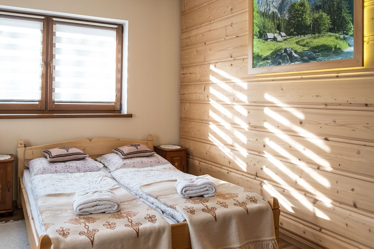 A double bed with floral-patterned bedding, two bedside tables, a window with blinds, and a framed landscape painting on the wall.