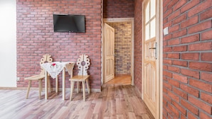 Una habitación con pared de ladrillo, un televisor de pantalla plana sobre una mesa con dos sillas, y una puerta de madera que conduce a otra habitación.