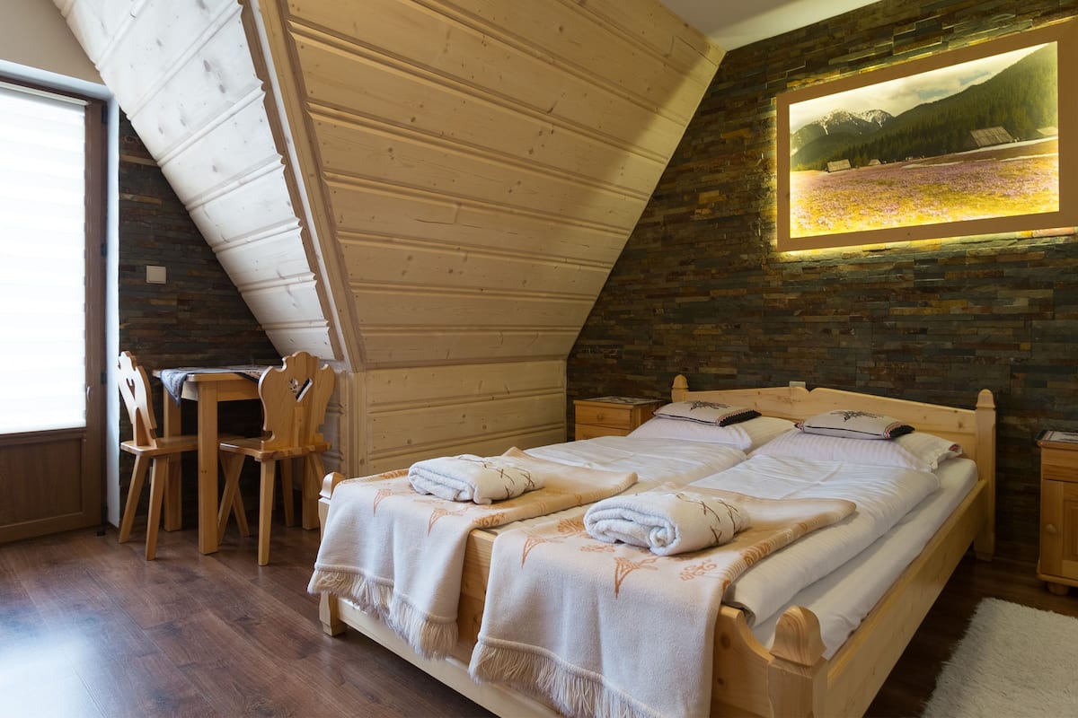 A bedroom with a sloped ceiling, a bed with white linens, a wooden nightstand, a chair, and a framed picture on the wall.