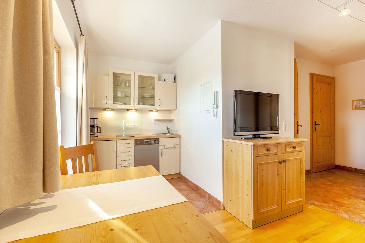 Cocina compacta con armarios de madera, lavavajillas integrado y un televisor sobre un mueble de madera.