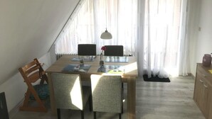 Zona pranzo con tavolo in legno, quattro sedie, lampada a sospensione e vista attraverso tende leggere.