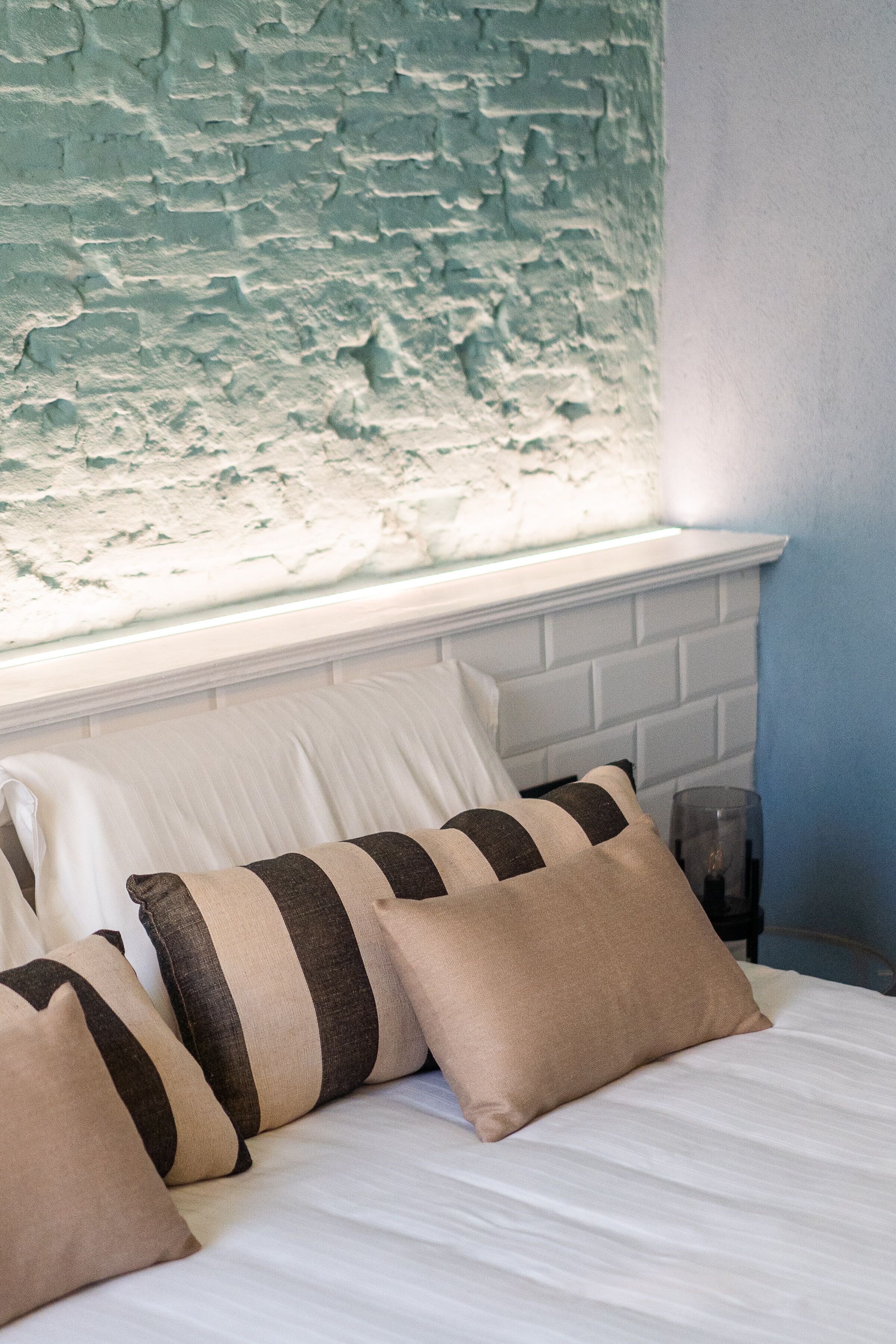 A bed with striped pillows and a white headboard in a room with a textured blue wall and a window above.