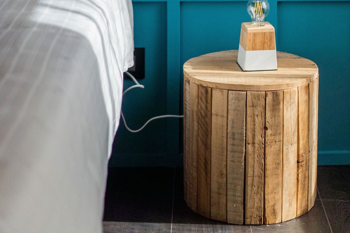 A bedroom with a bed, a wooden side table, and a wall-mounted light switch.