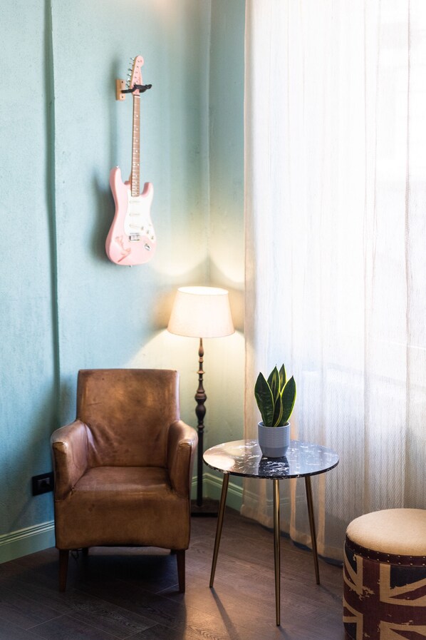 Una habitación con un sillón de cuero marrón, una mesita redonda con una lámpara y una planta en maceta, y una guitarra eléctrica rosa en la pared.