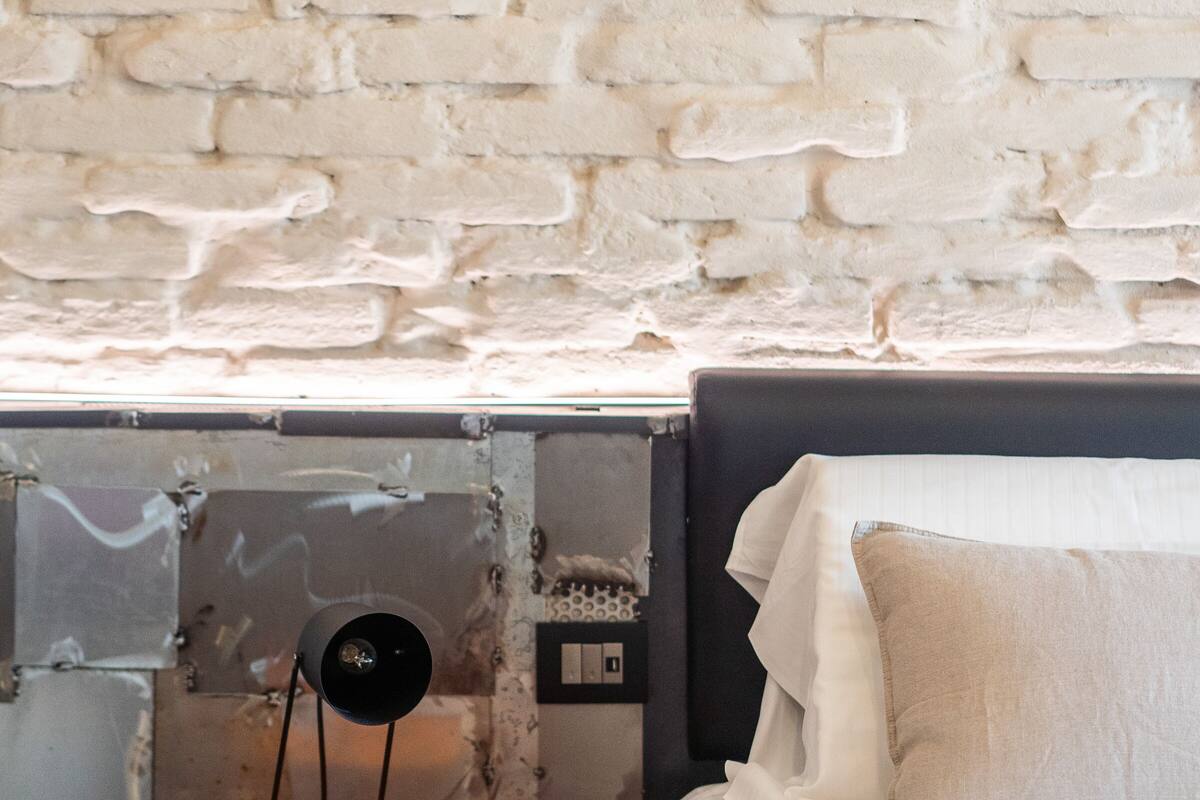 A bed with white bedding and a grey headboard, a white brick wall, and an orange cylindrical object with three drawers.