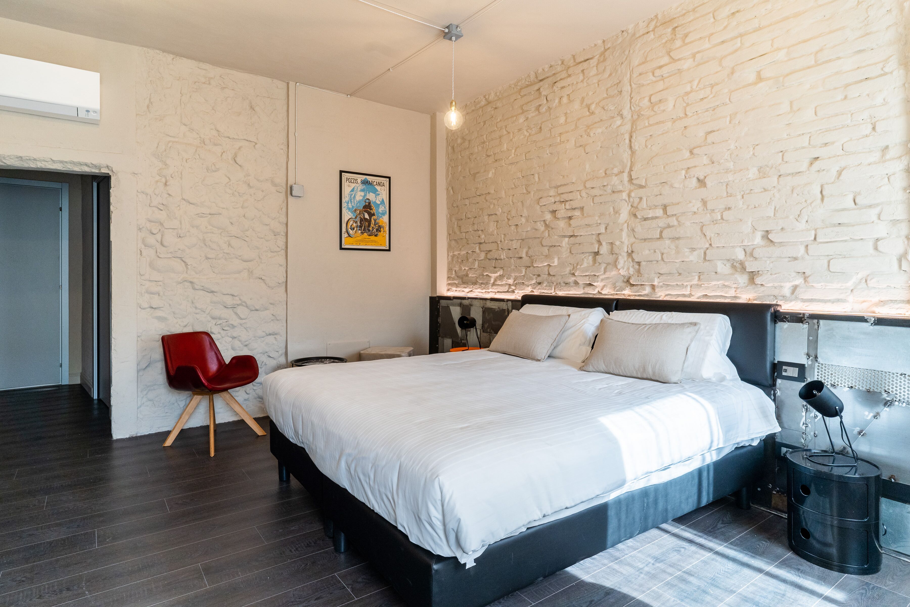 A modern bedroom with a large bed, a red chair, a white brick wall, and a poster on the wall.