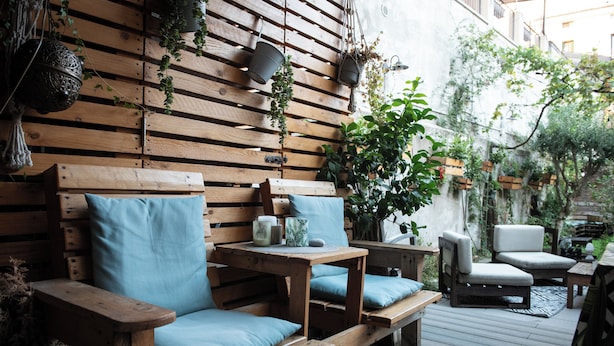 Eine Holzterrasse mit einer Couch und Stühlen, umgeben von Topfpflanzen und hängenden Pflanzgefäßen.