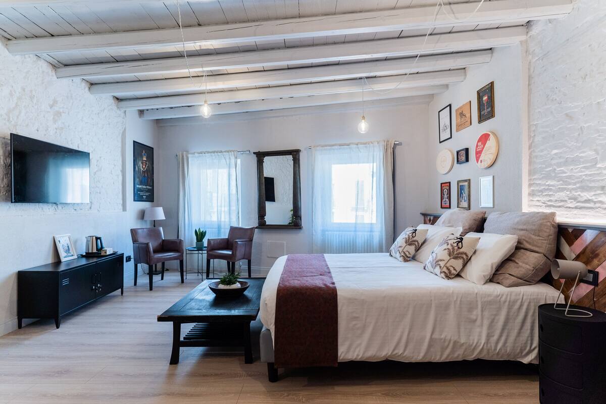 A spacious room with a large bed, a desk, and a chair. There is a television mounted on the wall and a window with curtains.