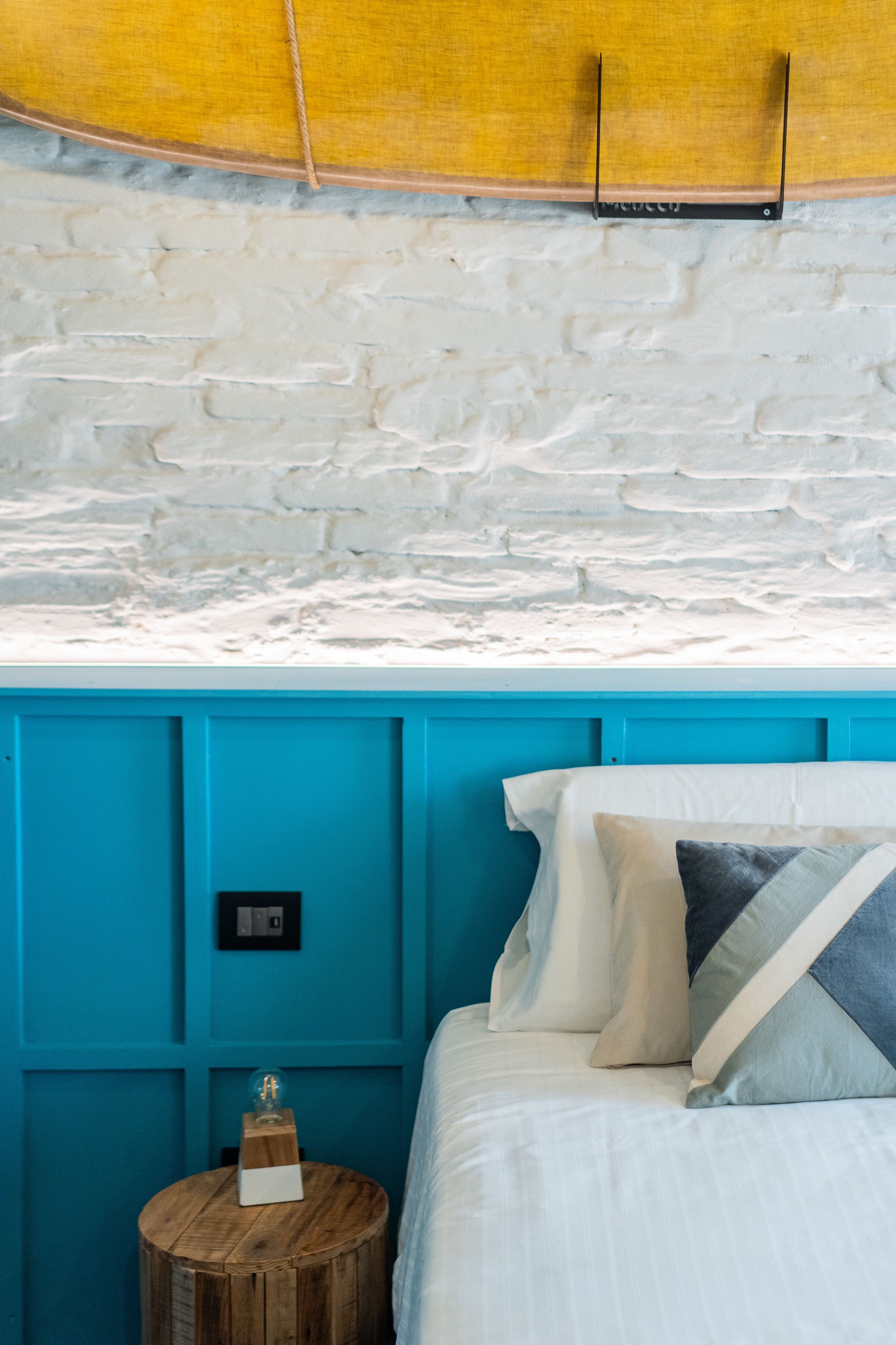 A bedroom with a white brick wall, a turquoise headboard, and a wooden nightstand with a lamp.
