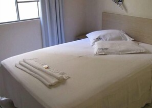 A neatly made bed with white linens and pillows, a headboard, and a window with curtains. - Hotel Jóia (Cascavel)