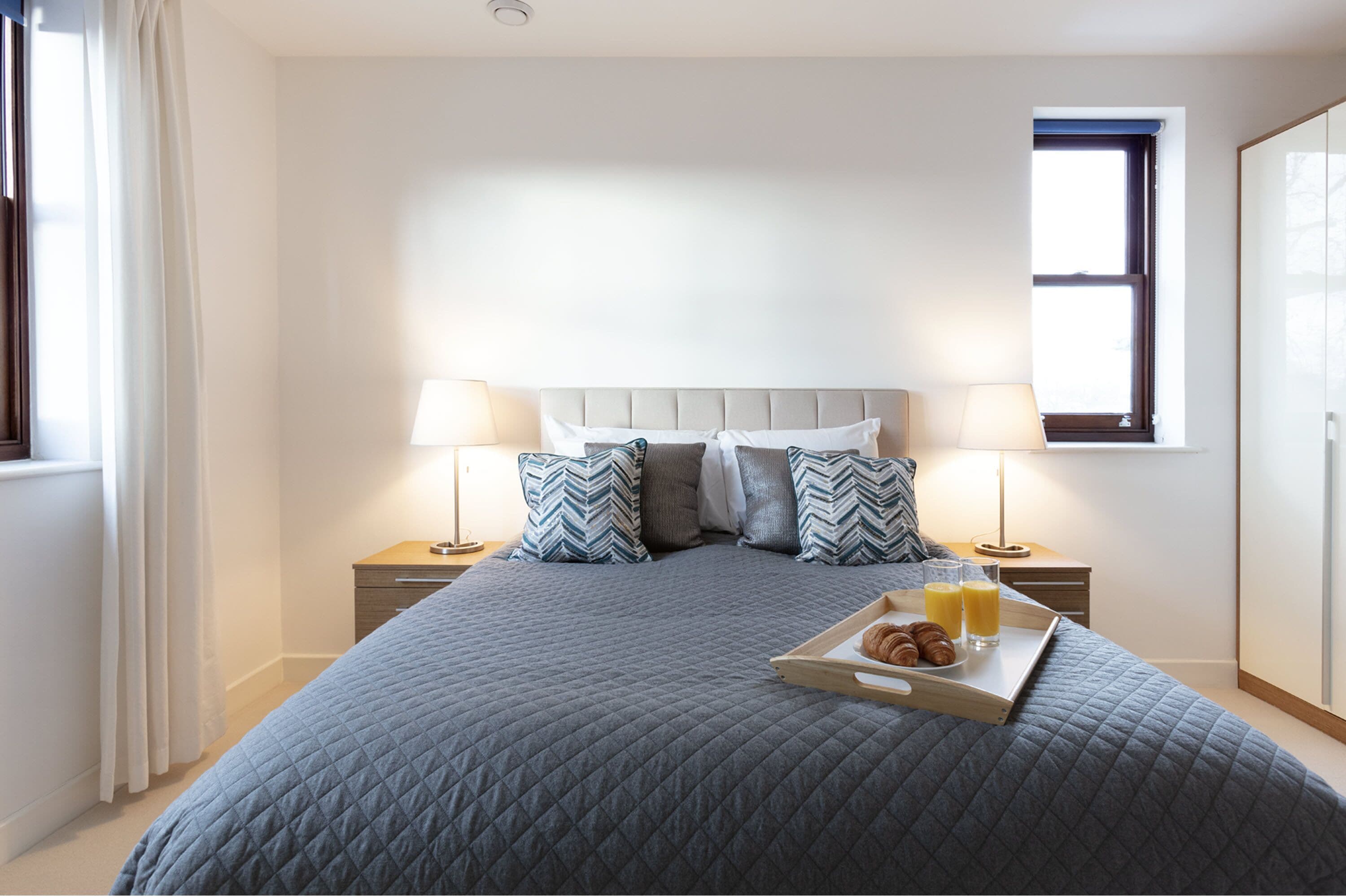 A neatly made bed with a grey quilt, two bedside tables with lamps, a tray with croissants and orange juice, and a window with a view.