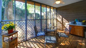 Alpendre coberto com ventilador de teto, mesa com cadeiras, pequena área de cozinha e vista para árvores através de grandes janelas.