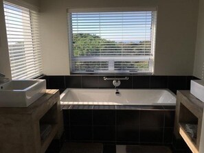 A modern bathroom with a freestanding bathtub, a window with blinds, and a view of greenery outside.
