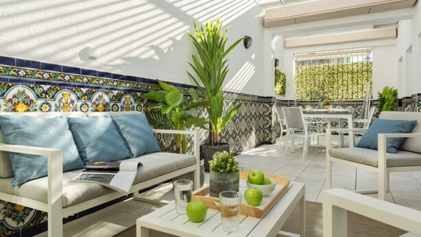 Um pátio ensolarado com sofá, cadeiras, uma mesa com maçãs e um copo, e um painel de azulejos.
