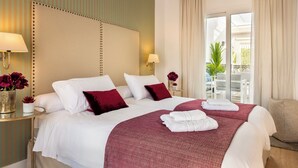 Quarto de hotel com cama grande, duas almofadas, mesa de cabeceira com abajur e vista para área externa com cadeiras e planta.