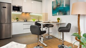 Uma cozinha moderna com armários brancos, uma geladeira de aço inoxidável, micro-ondas e uma área de jantar com uma mesa e dois bancos altos.