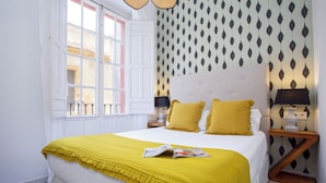 A neatly made bed with a yellow throw, a patterned wallpaper, and a window with white shutters. - Genteel Home Galera II (Seville)
