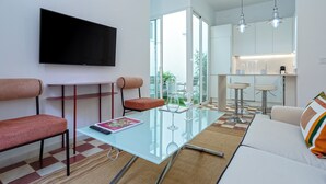 A modern living room with a glass coffee table, red upholstered chairs, a flat-screen TV, and a kitchen area with bar stools and pendant lights. - Genteel Home Galera II (Seville)
